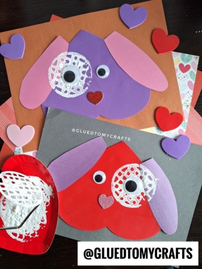 Two colorful Valentine dog crafts made from paper and googly eyes are displayed on colored backgrounds, surrounded by heart shapes. A small red plate with white paint hints at a fun potato masher stamped craft and painted doily. “@GLUEDTOMYCRAFTS” appears.