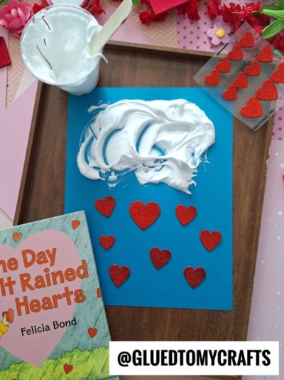 A craft setup inspired by the book "One Day It Rained Hearts" by Felicia Bond. The scene includes a blue paper with a white shaving cream cloud and red glitter hearts falling below it. The setup is arranged on a wooden tray with additional craft supplies like a cup of shaving cream, a spoon, and heart stickers.