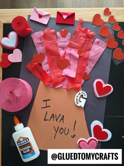 A handmade Valentine’s Day craft of a volcano made from brown, red, and pink paper, with hearts and “lava” erupting. Beside it are glue, heart decorations, and a tag reading “I Lava You!” with @GLUEDTOMYCRAFTS.