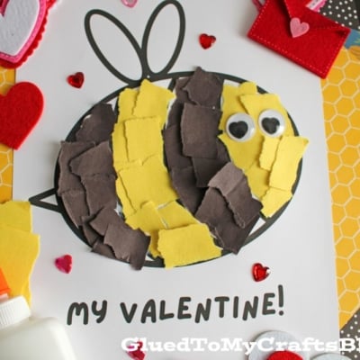 A vibrant Valentine's Day craft showcasing a torn-paper bumblebee with googly eyes on a white sheet, accompanied by the text "MY VALENTINE!" The scene is decorated with felt hearts, heart-shaped gems, and patterned papers, with a glue bottle and crafting supplies adding to the creative atmosphere.