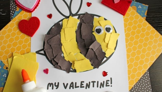 A vibrant Valentine's Day craft showcasing a torn-paper bumblebee with googly eyes on a white sheet, accompanied by the text "MY VALENTINE!" The scene is decorated with felt hearts, heart-shaped gems, and patterned papers, with a glue bottle and crafting supplies adding to the creative atmosphere.
