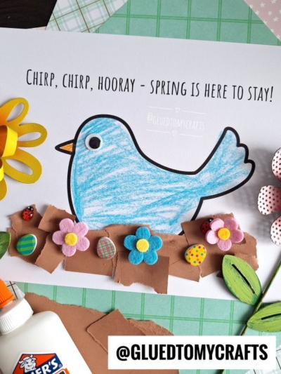 A handmade spring craft featuring a blue paper bird in a torn paper bird nest, colorful felt flowers, paper leaves, and ladybug decorations on a white sheet with the text Chirp, chirp, hooray - spring is here to stay!.
