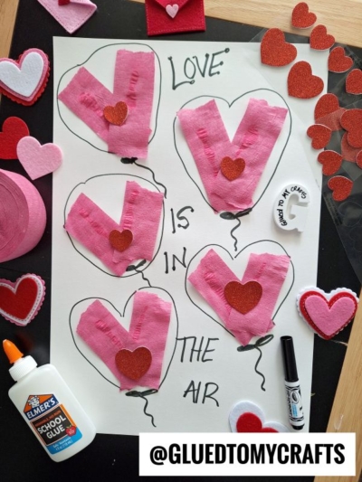 A Valentine’s Day craft featuring five pink paper heart balloons with red glitter hearts, surrounded by heart cutouts, marker, glue, and a sticker reading “GLUED TO MY CRAFTS.” The text says Love is in the air.