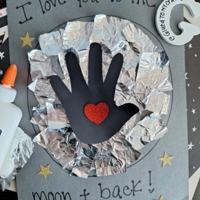 A handmade card features a black paper handprint with a red heart sticker in the center, surrounded by crumpled foil resembling the moon. The card says, “I love you to the moon & back!” with gold star stickers.