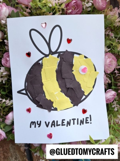A handmade Valentine’s card features a torn paper bee crafted from yellow and black pieces on white paper, with the words “MY VALENTINE!” Decorative heart gems surround the card, set among pink flowers.