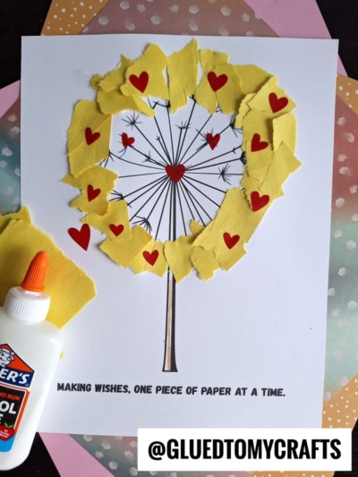 A torn paper dandelion craft made with yellow bits, red heart shapes, and black marker lines on white paper. Elmer’s glue bottle is nearby. Text below reads: MAKING WISHES, ONE PIECE OF PAPER AT A TIME.