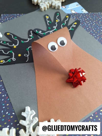 A handmade reindeer craft with brown paper, googly eyes, a shiny red bow for a nose, and black antlers decorated with colorful lights, displayed on a dark square background with snowflake decorations.