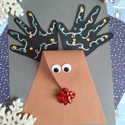 A handmade reindeer craft with black paper handprints as antlers, decorated with dots and squiggles, a brown triangular face with googly eyes and a red bow nose, surrounded by paper snowflakes.