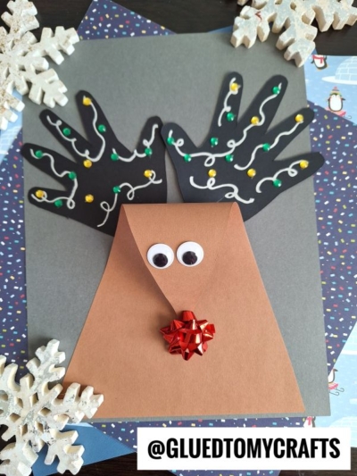 A handmade reindeer craft with black paper handprints as antlers, decorated with dots and squiggles, a brown triangular face with googly eyes and a red bow nose, surrounded by paper snowflakes.
