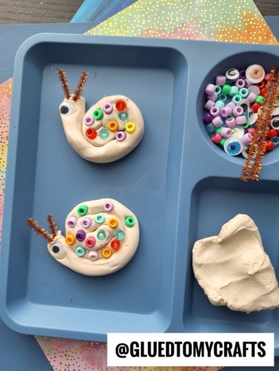 Two snail crafts made from white modeling clay, colorful beads for shells, googly eyes, and brown pipe cleaners for antennae are displayed in a blue tray with craft supplies and the handle @GLUEDTOMYCRAFTS.