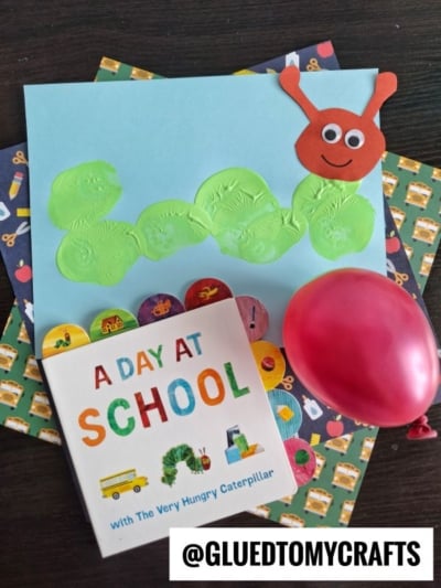 A children’s HUNGRY CATERPILLAR CRAFT project featuring a green caterpillar and red caterpillar head on blue paper, a red balloon, the book “A Day at School,” and colorful school-themed patterned paper.