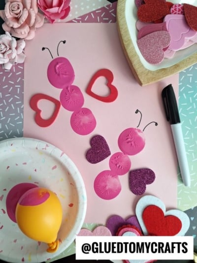 A pink butterfly paper craft made with pink balloon paint stamps for the body and foam hearts for wings. Art supplies and heart shapes are scattered around—perfect for INSECT CRAFT IDEAS. @GLUEDTOMYCRAFTS at the bottom.