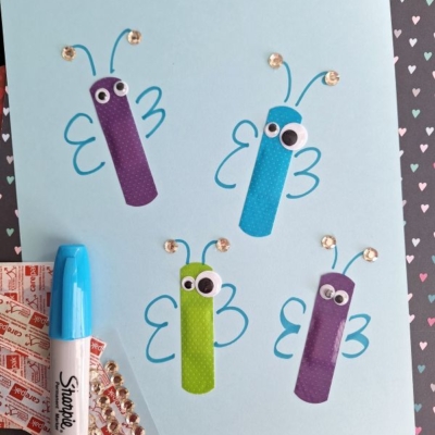 A light blue paper displays a Band-Aid Butterfly Craft featuring four butterflies made from colored bandages, googly eyes, hand-drawn blue wings, and gem decorations. A blue marker and gem stickers are nearby. The “@GLUEDTOMYCRAFTS” handle is at the bottom.