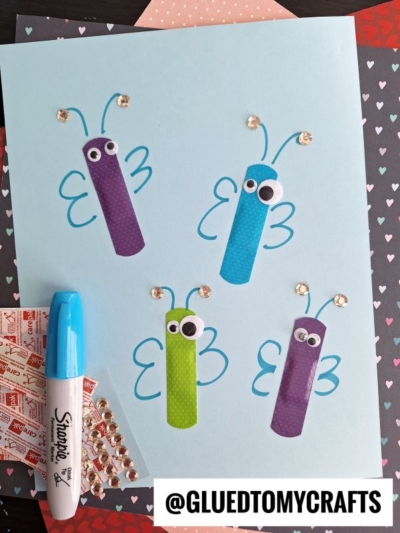 A light blue paper displays a Band-Aid Butterfly Craft featuring four butterflies made from colored bandages, googly eyes, hand-drawn blue wings, and gem decorations. A blue marker and gem stickers are nearby. The “@GLUEDTOMYCRAFTS” handle is at the bottom.