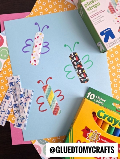 A blue sheet of paper showcases a charming Band-Aid Butterfly Craft—four butterflies made from colorful patterned bandaids and marker wings, surrounded by crayons, plastic strips, and patterned paper. @GLUEDTOMYCRAFTS is written at the bottom.