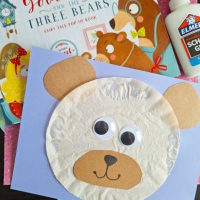 A children’s craft project of a bear’s face made with tissue paper, construction paper, and googly eyes sits on a desk alongside a Goldilocks and the Three Bears book, glue, colorful sheets of paper, and other fun coffee filter crafts for kids.