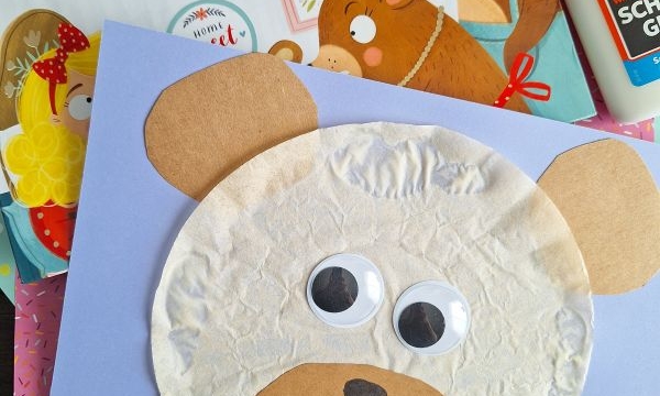 A children’s craft project of a bear’s face made with tissue paper, construction paper, and googly eyes sits on a desk alongside a Goldilocks and the Three Bears book, glue, colorful sheets of paper, and other fun coffee filter crafts for kids.