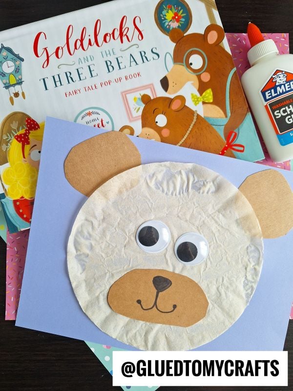A children’s craft project of a bear’s face made with tissue paper, construction paper, and googly eyes sits on a desk alongside a Goldilocks and the Three Bears book, glue, colorful sheets of paper, and other fun coffee filter crafts for kids.