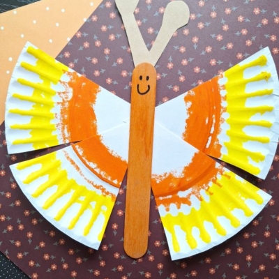 A butterfly craft made from painted paper plate pieces for wings, colored orange and yellow, and a wooden craft stick for the body with a smiley face, displayed on patterned paper. Text: @GLUEDTOMYCRAFTS.