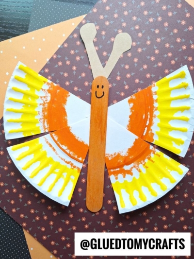 A butterfly craft made from painted paper plate pieces for wings, colored orange and yellow, and a wooden craft stick for the body with a smiley face, displayed on patterned paper. Text: @GLUEDTOMYCRAFTS.