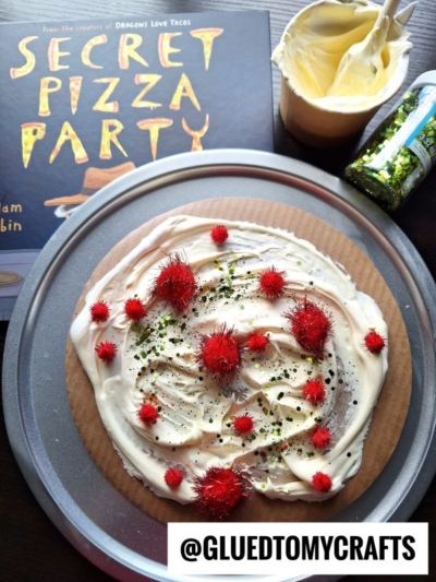 A pie plate with a pizza craft made from white frosting, red pom-poms, and green sprinkles—perfect for puffy paint crafts fans. Nearby are a frosting tub, green sprinkles jar, and the Secret Pizza Party book. @GLUEDTOMYCRAFTS is labeled.
