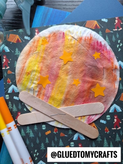 A colorful craft depicting a campfire made with a coffee filter colored in red, orange, and yellow, five yellow star stickers, and two craft sticks on a camping-themed background. Markers and a spray bottle are nearby.