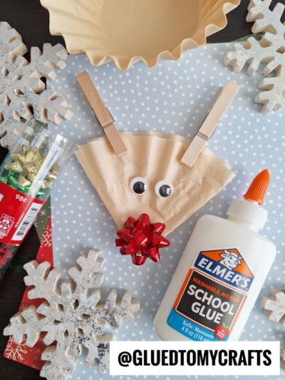 A reindeer craft made from a coffee filter, clothespins for antlers, googly eyes, and a red bow nose is shown on blue snowflake paper—an adorable idea for coffee filter crafts for kids, with glue, mini bows, and large snowflake decorations nearby.