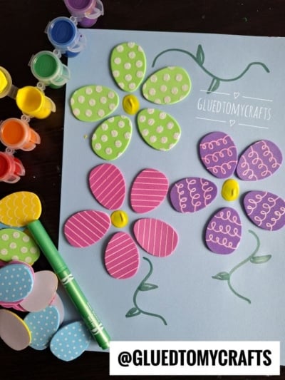 A craft project with colorful foam petals forming two flowers on blue paper, decorated with white marker designs. Paint pots and a green marker are nearby. @GLUEDTOMYCRAFTS is written at the bottom.