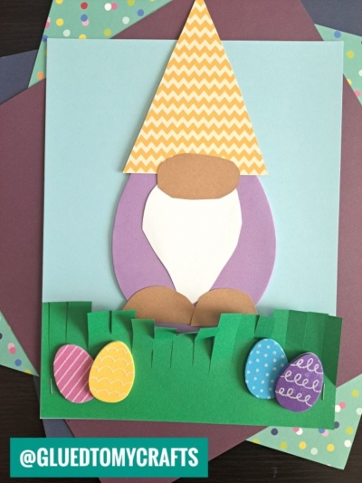 A paper craft of an Easter gnome with a yellow patterned hat, white beard, and brown nose, standing in green grass with three colorful Easter eggs on a pastel background.