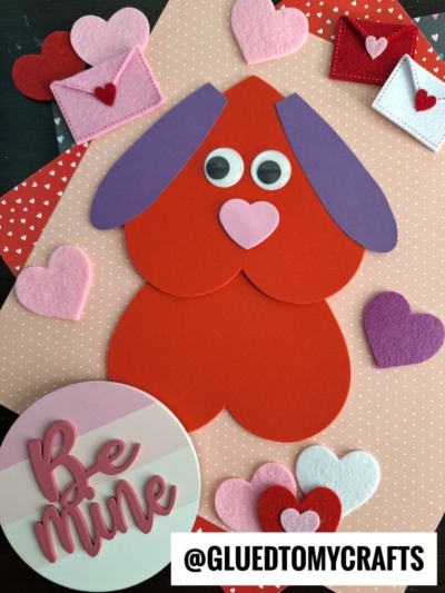 A heart shaped Valentine’s Day craft featuring a dog made from red, purple, and pink paper hearts, surrounded by felt and craft foam heart crafts, mini envelopes, and a “Be Mine” sign on a polka-dot background—perfect for valentine art project ideas.