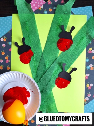 A spring INSECT CRAFT IDEA features ladybugs made from red-painted egg cartons with black paper heads and antennae, displayed on green paper “stems” and surrounded by colorful patterned paper. A paint-filled plate and egg are nearby.