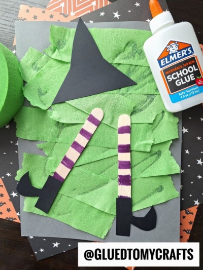 A Halloween crepe paper craft idea features a witch’s hat and legs made from paper and popsicle sticks, with green crepe paper for the body, black shoes, Elmer’s school glue nearby, and the Instagram handle @gluedtomycrafts visible.