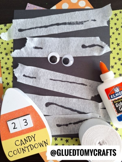 A Halloween craft scene with a black paper mummy made using torn white crepe paper strips, googly eyes, Elmer’s glue, a roll of crepe paper, and a Candy Countdown sign—perfect inspiration for crepe paper craft ideas.