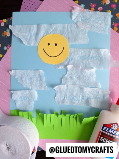 A cheerful kids’ craft featuring a paper sun with a smiley face, crepe paper clouds, and green paper grass on a blue background—perfect for those seeking fun crepe paper craft ideas. Elmer’s glue and rolls of crepe paper are nearby. @GLUEDTOMYCRAFTS at the bottom.