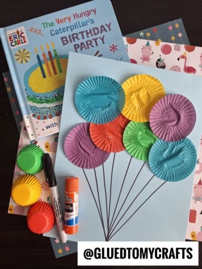 A birthday craft using colorful mini cupcake liners arranged as balloon shapes on blue paper—perfect for cupcake liner card crafts—with markers, glue, a book titled The Very Hungry Caterpillar's Birthday Party, and patterned paper.