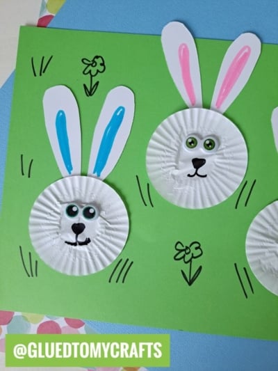 Two bunny faces made from white cupcake liners with googly eyes, drawn noses and mouths, and paper ears decorated with blue and pink create a fun cupcake liner card crafts project on green paper with hand-drawn flowers, grass, and a @GLUEDTOMYCRAFTS label.