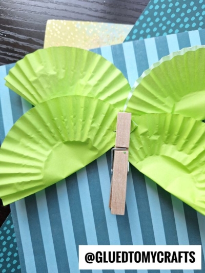 A butterfly craft made with green cupcake liners for wings and a wooden clothespin for the body, displayed on striped blue paper. The text @GLUEDTOMYCRAFTS appears at the bottom.