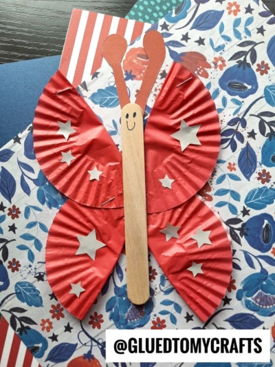 A butterfly craft made with red cupcake liners for wings, decorated with silver star stickers, and a wooden popsicle stick for the body, placed on floral-patterned paper. @GLUEDTOMYCRAFTS is written at the bottom.