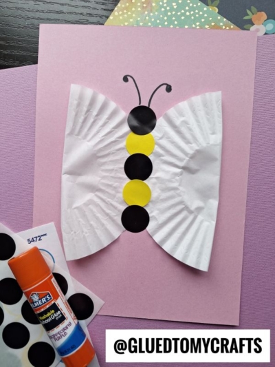 A butterfly craft made from a cupcake liner, black and yellow paper circles, and black antennae, on a pink background with craft supplies nearby. @GLUEDTOMYCRAFTS is printed at the bottom.