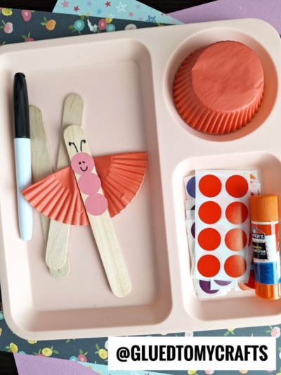 A craft tray with materials for coding label crafts: craft sticks, a black marker, orange cupcake liners, glue stick, red circle stickers, and a butterfly made from these items. @GLUEDTOMYCRAFTS is noted at the bottom.