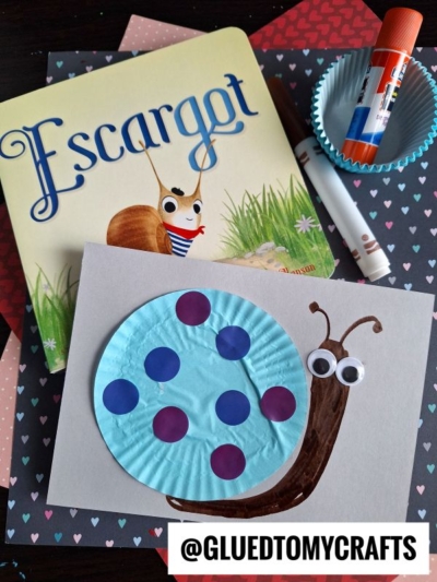 A snail craft made from a blue cupcake liner with purple dots, googly eyes, and marker details sits on gray paper. Nearby are the book Escargot, a glue stick, a marker, scrapbook paper, and coding label crafts supplies.
