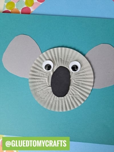 A koala face cupcake liner card craft made from a gray cupcake liner, with two large googly eyes, a black oval nose, and gray paper ears on a blue background. A label at the bottom reads @GLUEDTOMYCRAFTS.