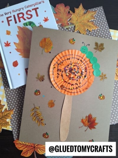 A children’s HUNGRY CATERPILLAR CRAFT features a smiling caterpillar made from green circles on an orange paper swirl, glued to brown paper with leaf stickers. Nearby is a copy of The Very Hungry Caterpillar’s First Fall book.