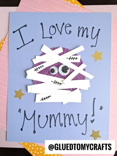 Handmade card on a blue background with the text “I Love my Mummy!” A mummy face made of white paper strips and googly eyes is in the center, inspired by cupcake liner card crafts, with gold stars and @GLUEDTOMYCRAFTS at the bottom.