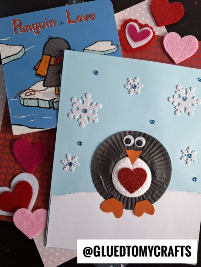 A handmade penguin Valentine’s card with a cupcake liner body, heart and snowflake decorations, beside the book Penguin in Love and felt heart shapes. The handle @GLUEDTOMYCRAFTS is visible at the bottom.