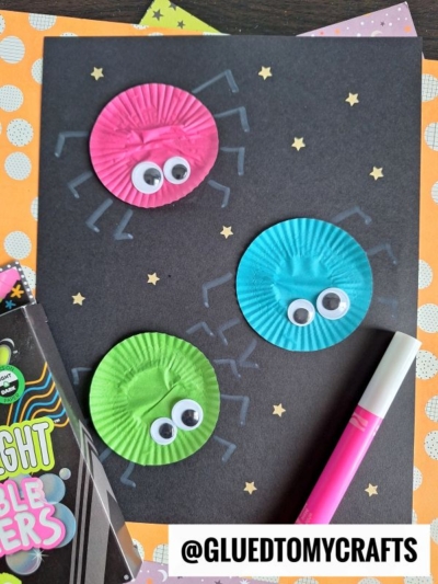 A black paper showcases three spider crafts made from pink, blue, and green cupcake liners with googly eyes and drawn legs—perfect for INSECT CRAFT IDEAS. Halloween-themed items, orange polka dot paper, and a pink marker are nearby.
