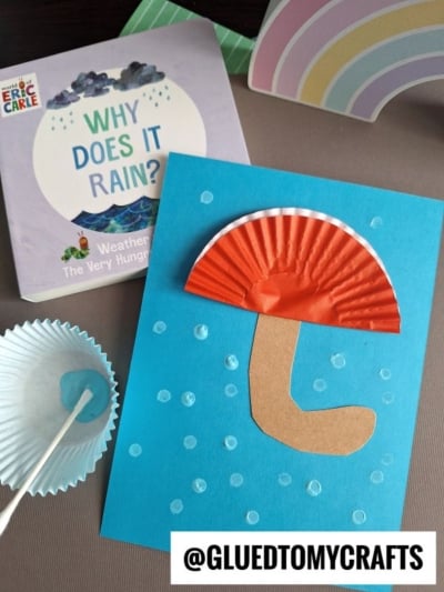 A children's weather-themed craft featuring a cupcake liner umbrella on blue paper, inspired by the book Why Does It Rain? by Eric Carle, with a striped paper rainbow in the background.