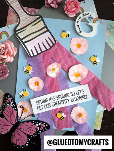 A spring-themed craft with tissue paper strips, paper flowers, bees, a paintbrush cutout, and a cute Fingerprint Daisy on blue paper. Text reads “Spring has sprung, so let’s get our creativity blooming!” and “@GLUEDTOMYCRAFTS.”.