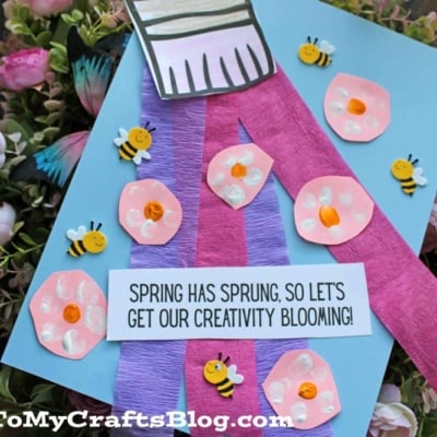 A spring-themed craft featuring pink and purple paper flowers, small decorative bees, and a “Spring has sprung, so let’s get our creativity blooming!” sign on a blue background—perfect for pairing with Spring Printables.