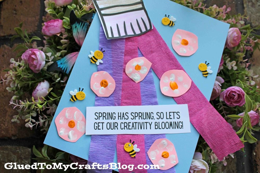 A spring-themed craft featuring pink and purple paper flowers, small decorative bees, and a “Spring has sprung, so let’s get our creativity blooming!” sign on a blue background—perfect for pairing with Spring Printables.